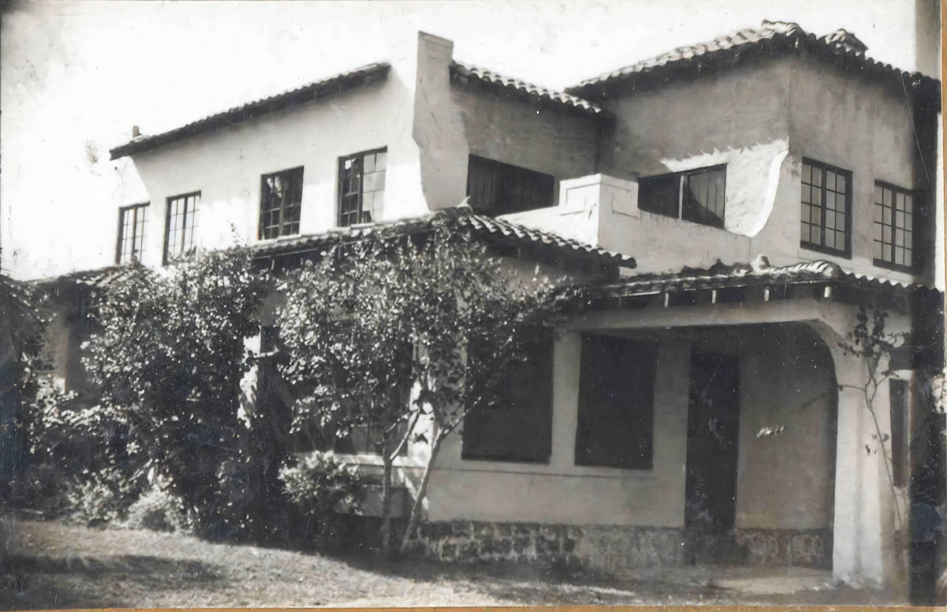 This image showcases a vintage, two-story house with Spanish architectural influences, featuring stucco walls, terracotta roof tiles, and a courtyard with lush greenery. The photo has been enhanced with the 'Upscale' tool, which preserves the original image's authenticity while improving its clarity and detail, highlighting the texture of the stucco and the vibrant foliage of the plants.